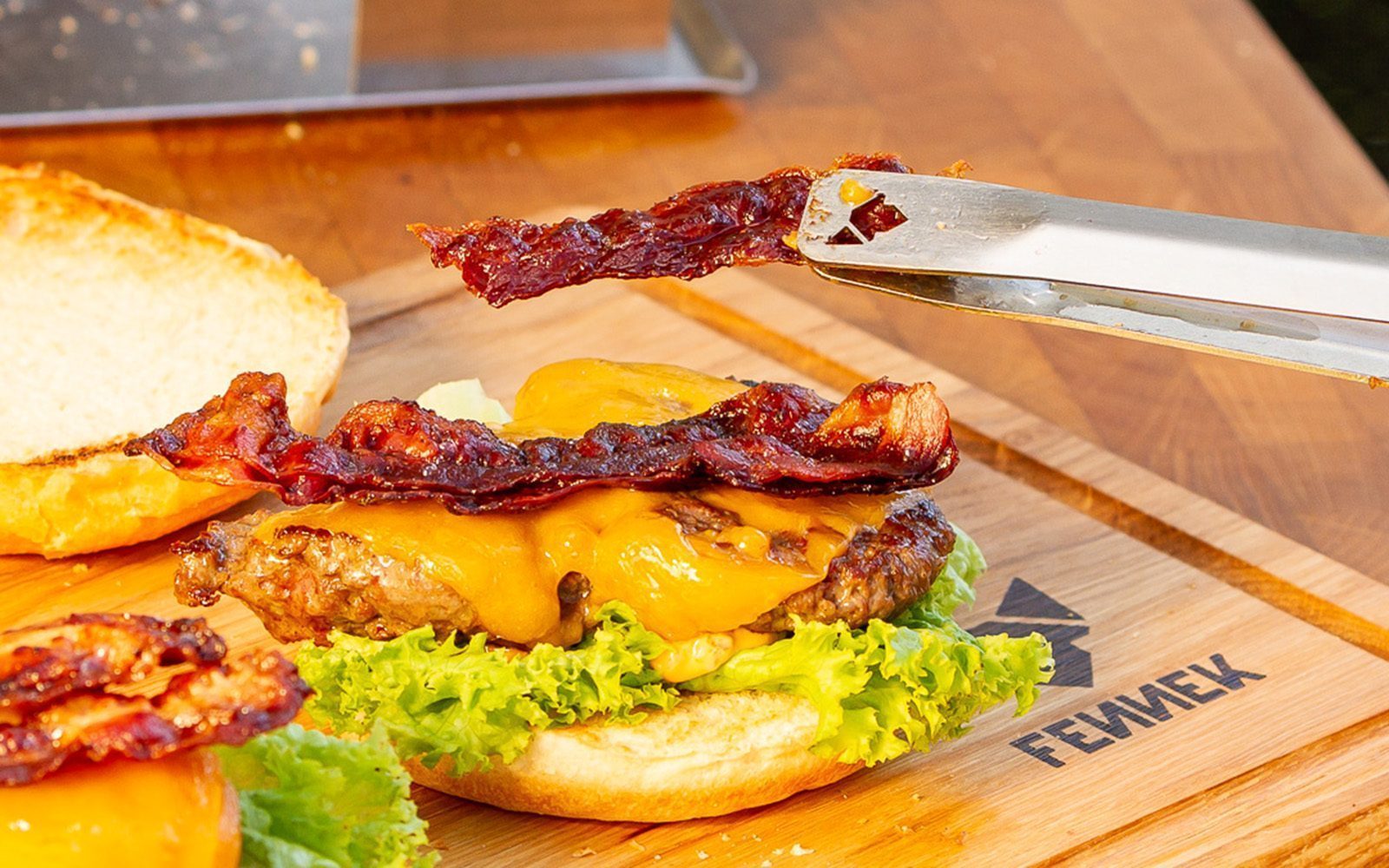 Mit Grillzange wird ein Stück knusprig gebratener Bacon auf einen Hamburger gelegt.