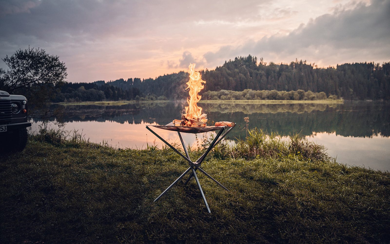 Das in der Natur aufgestellte Feuernetz im Einsatz mit brennendem Holz darauf und einer stimmungsvollen Aussicht auf ein stilles Gewässer mit Spiegelung der dichtbewachsenen Landschaft im Hintergrund.