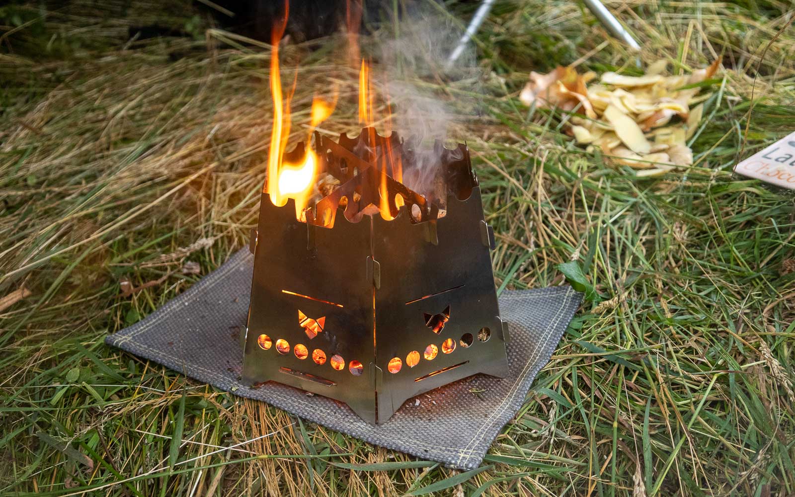 Der Hobokocher FENNEK Basic mit Erweiterungsset mit Flammen darin auf einer Hitzeschutzmatte im Gras