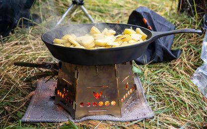 Der Hobokocher FENNEK Basic mit Erweiterungsset in Aktion beim Zubereiten von Bratkartoffeln mit einer schwarzen Gußpfanne auf einer Wiese.