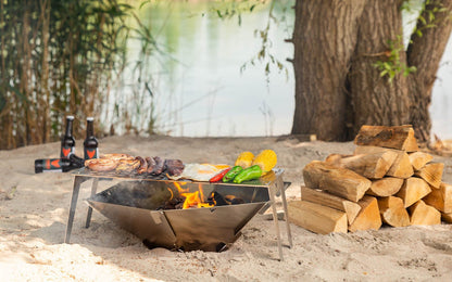 Feuerschale Hexagon mit brennendem Holz am Sandstrand mit Grillrost 4Fire und gegrilltem Fleisch, Spiegeleier und buntem Gemüse darauf.
