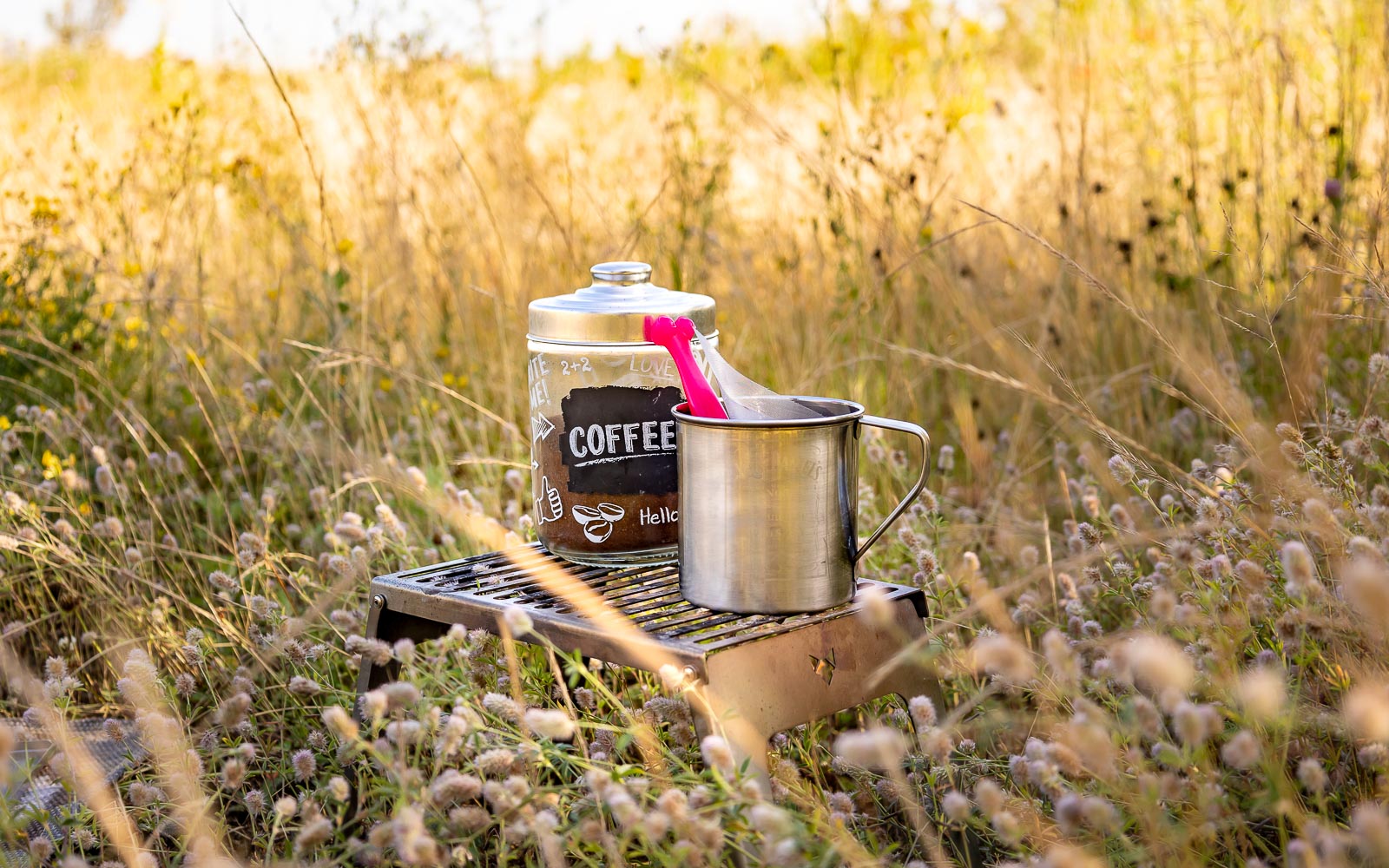 Klapprost als Tisch auf einer wilden Sommerwiese genutzt für einen Behälter mit Kaffeepulver darin und eine Edelstahl-Tasse