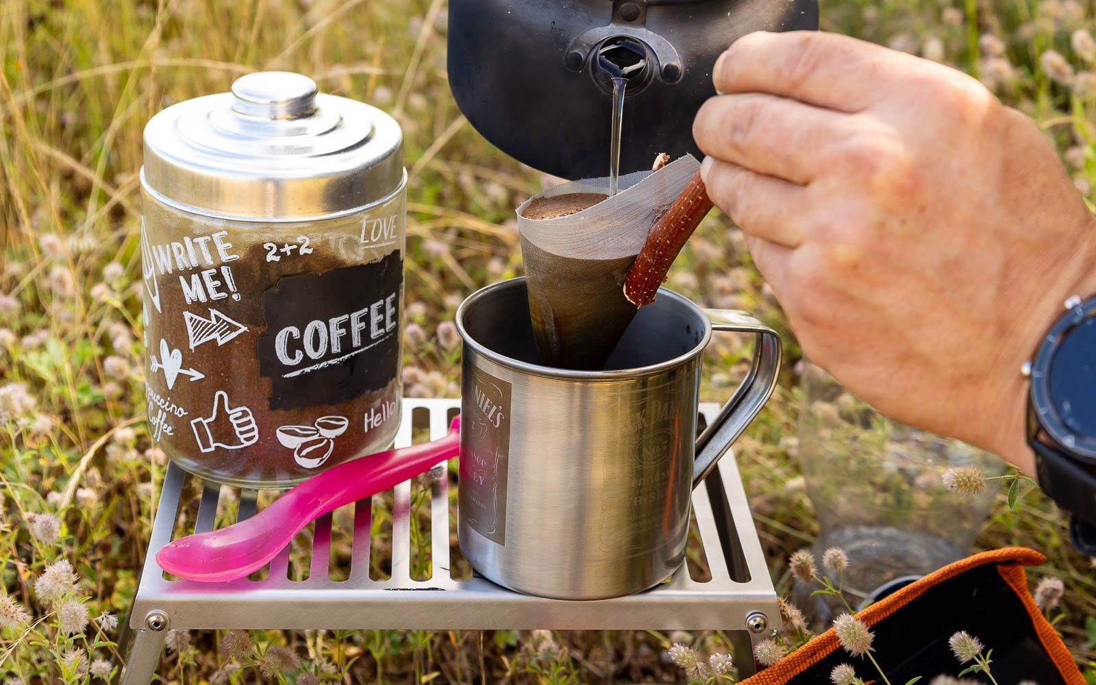 Kaffee wird mit heißem Wasser aus einem schwarzen Wasserkessel in einem Kaffeefilter aufgegossen der in eine Edelstahltasse gehalten wird die, wie auch der Behälter mit dem Kaffeepulver auf dem Klapprost abgestellt ist.