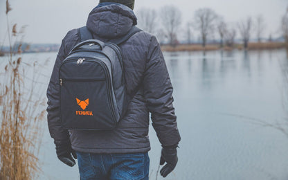 Rucksack mit FENNEK Logo-Stick auf dem Rücken getragen vor einem Gewässer an einem trüben Tag im Herbst/Winter