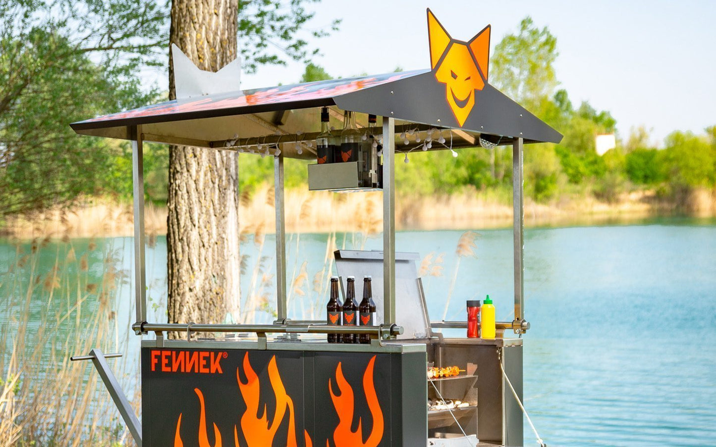 FENNEK Bollerwagen mit Deichsel vor einem See bei sonnigem Wetter und eingebauten Holzkohlegrill.