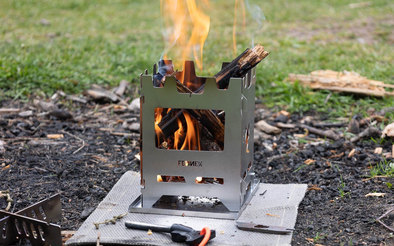 Edelstahl Hobokocher auf einer Hitzschutzmatte in der Natur im Einsatz mit Blick durch die Frontöffnung auf das brennenden Holz, davor liegt ein Feuerstahl