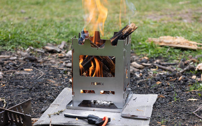 Brennendes Holz in einem Hobokocher der auf der Multischale steht und darunter eine Brandschutzmatte um den Waldboden zu schützen. Davor liegt ein Feuerstahl., im Hintergrund grüner Rasen.