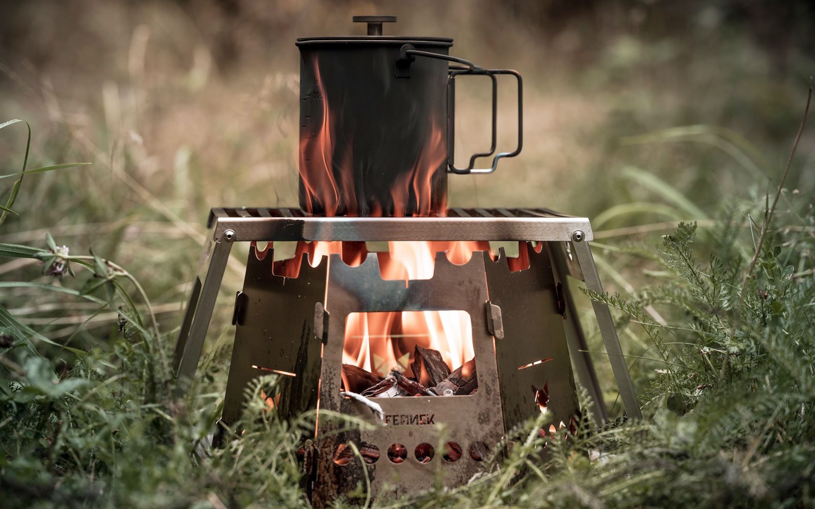 Outdoortasse auf einem Klapprost über einem Feuer das im Hobokocher mit Erweiterungs-Set brennt in der grünen Natur.