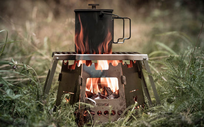 6-eckiger Hobokocher mit brennendem Holz darin steht in der grünen Natur und befeuert eine große Outdoor-Tasse die auf dem Klapprost steht.