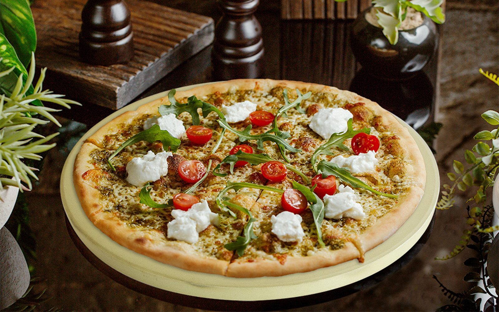 Pizzastein mit einer frisch gebackenen Pizza darauf die mit Käse, Rucolasalat und Tomaten belegt ist.