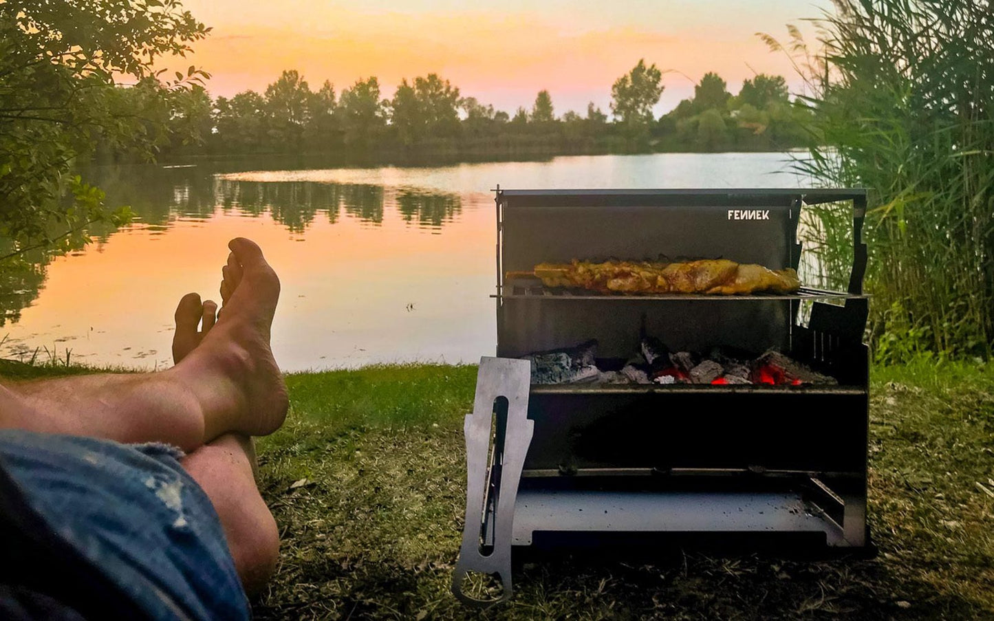 Fennek Grill mit glühender Kohle und Grillfleisch auf dem Rost bei Sonnenuntergang am See.