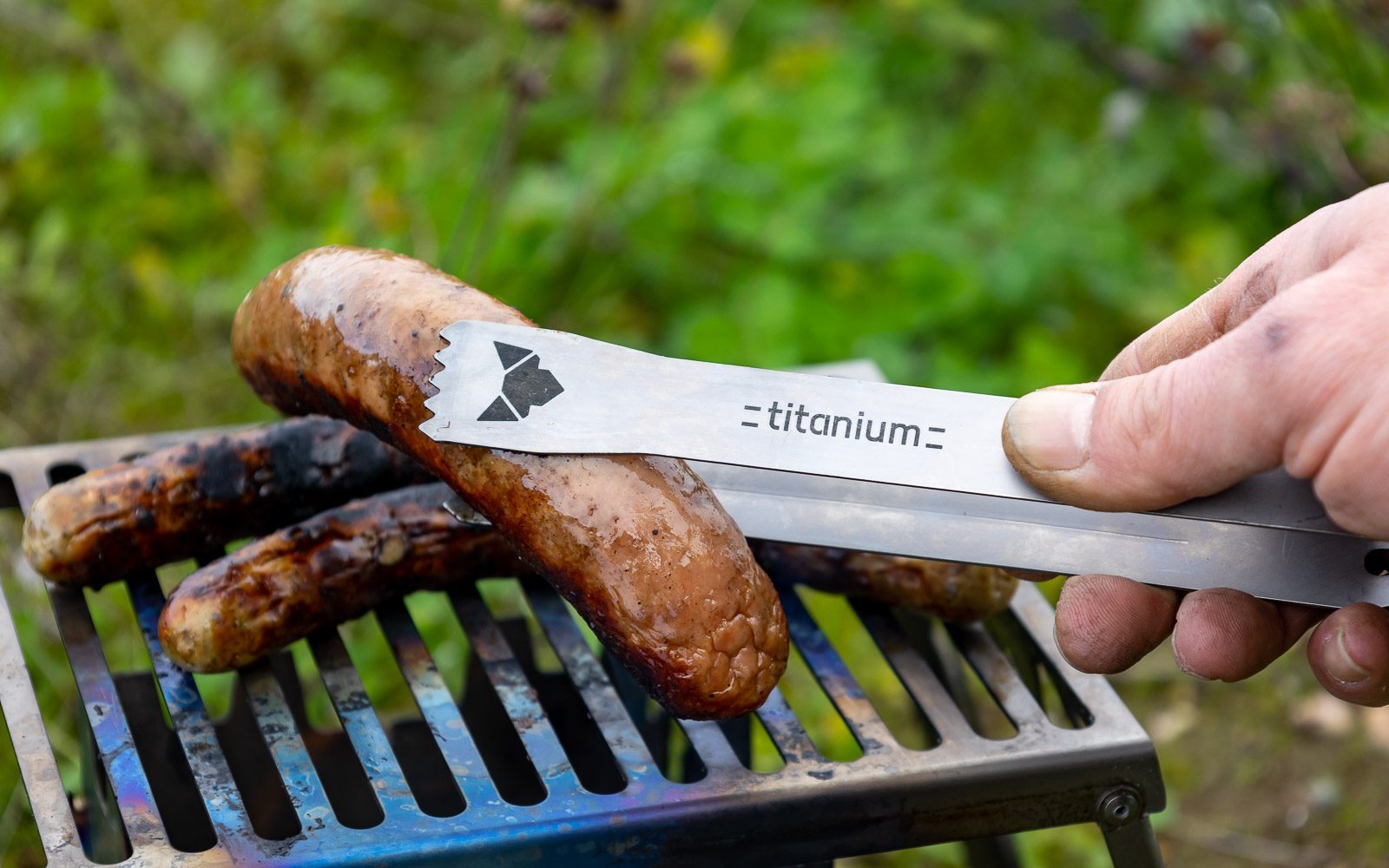 Kleine Grillzange aus Titan mit laser-graviertem FENNEK-Logo und Titanium-Schriftzug im Einsatz beim Greifen einer Bratwurst