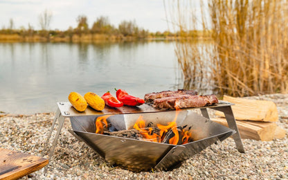 Feuerschale FENNEK Hexagon im Einsatz am Kiesstrand in Kombination mit dem Grillrost 4Fire auf dem Fleisch und Gemüse gegrillt wird. Im Hintergrund unscharf ein See und Schilf..