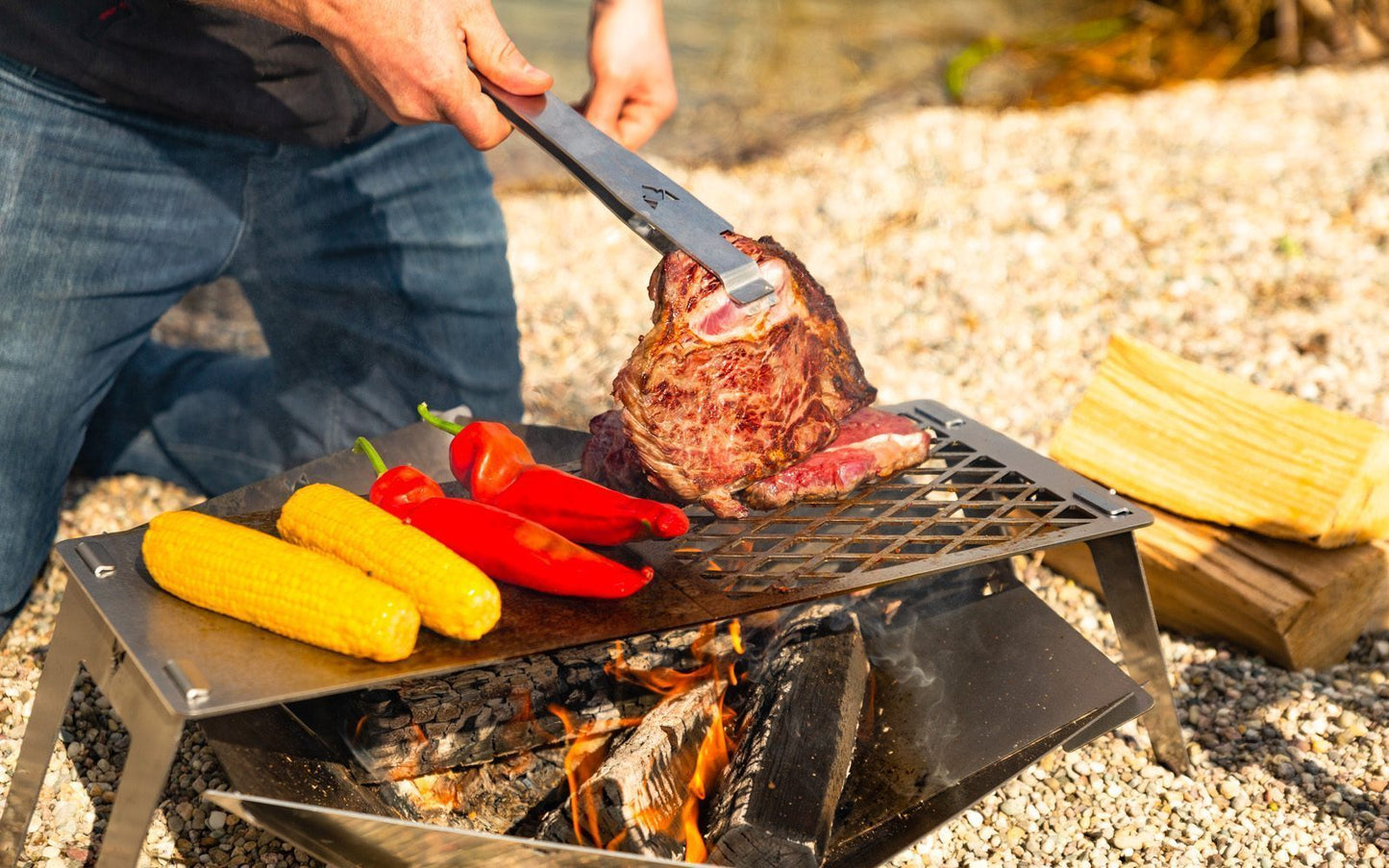 Grillzange wendet ein großes Steak auf der mobilen Grillplatte über der Feuerschale, Maiskolben und Spitzpaprika liegen auch auf dem Grill.