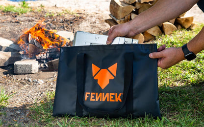 Einzelteile von Grillrost 4Fire werden aus der schwarzen Tarpaulin Henkeltasche mit FENNEK Logo-Aufdruck herausgezogen. Im Hintergrund brennt ein offenes Feuer.