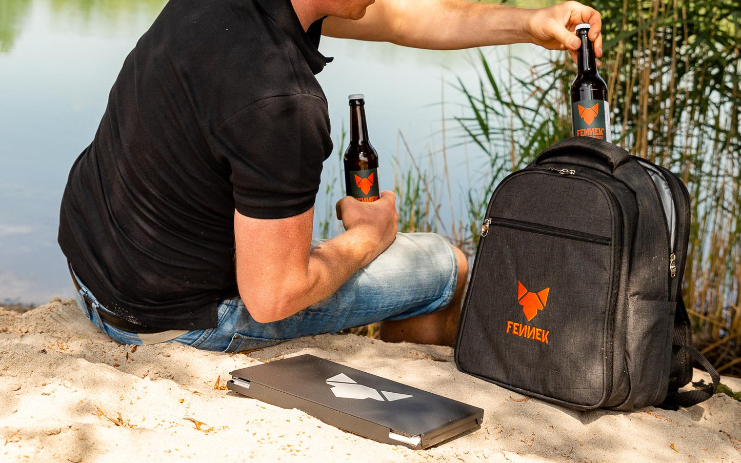 Rucksack mit FENNEK Logo-Applikation und FENNEK Grill in schwarzer Alubox liegen im Sand vor einem See und jemand holt 2 Flaschen aus dem Kühlfach.