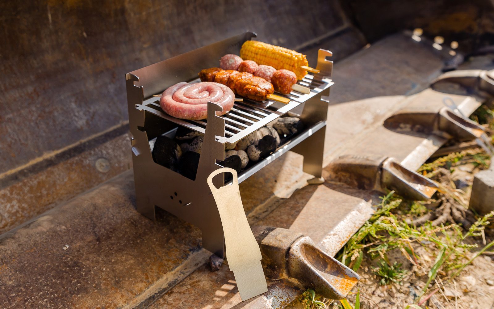 Mobiler Holzkohlegrill im Einsatz, steht in einer Baggerschaufel und grillt bei sonnigem Wetter verschiedenes Fleisch und einen Maiskolben.
