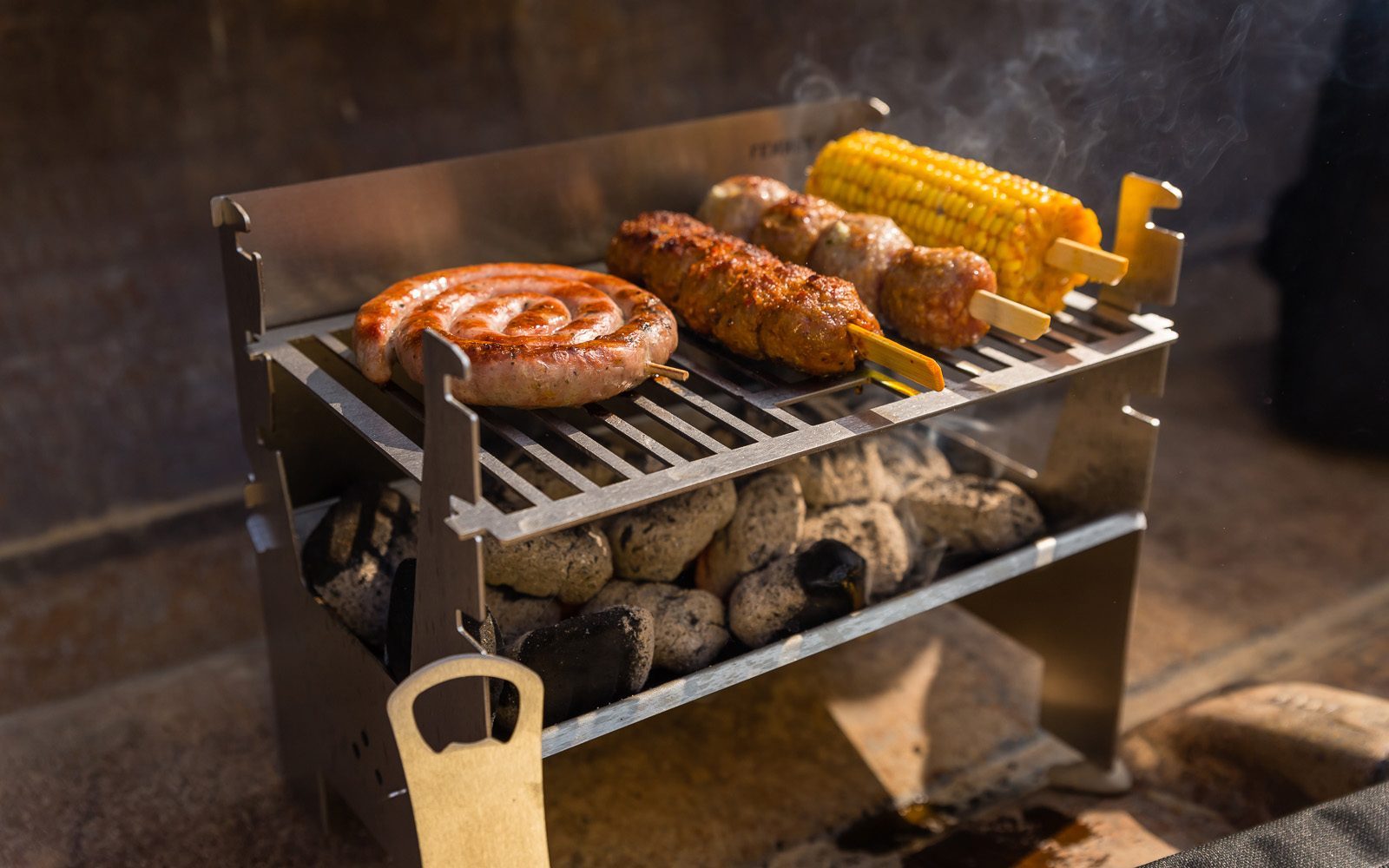 Lecker gebratene Bratwurstschnecke und weiteres Fleisch am Spieß und ein Maiskolben auf einem mobilen Holzkohlegrill aus Edelstahl.