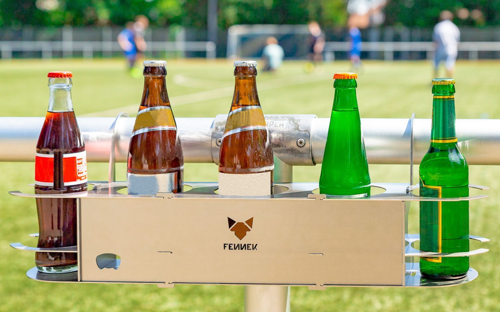 Fünf Glasflaschen in einem Getränkehalter aus Edelstahl mit FENNEK-Logo in der Mitte, befestigt am Fussballspielfeldrand. Im Hintergrund unscharf Fussballspieler.