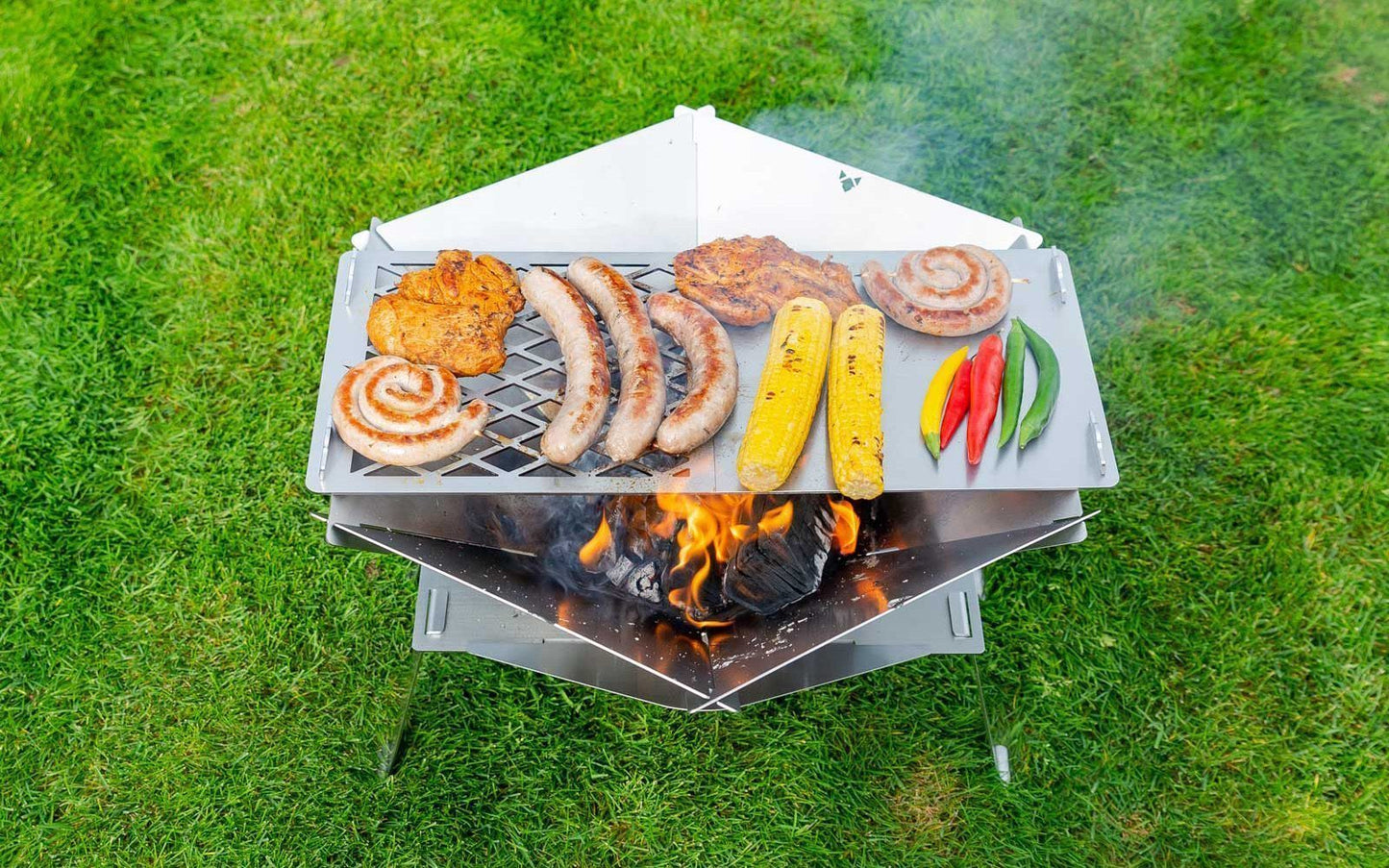 Fleisch und Gemüse wird auf einer zusammengesteckten Grillplatte über einer Feuerschale gegrillt die sich mit Erhöhung auf einem grünen Rasen befindet.
