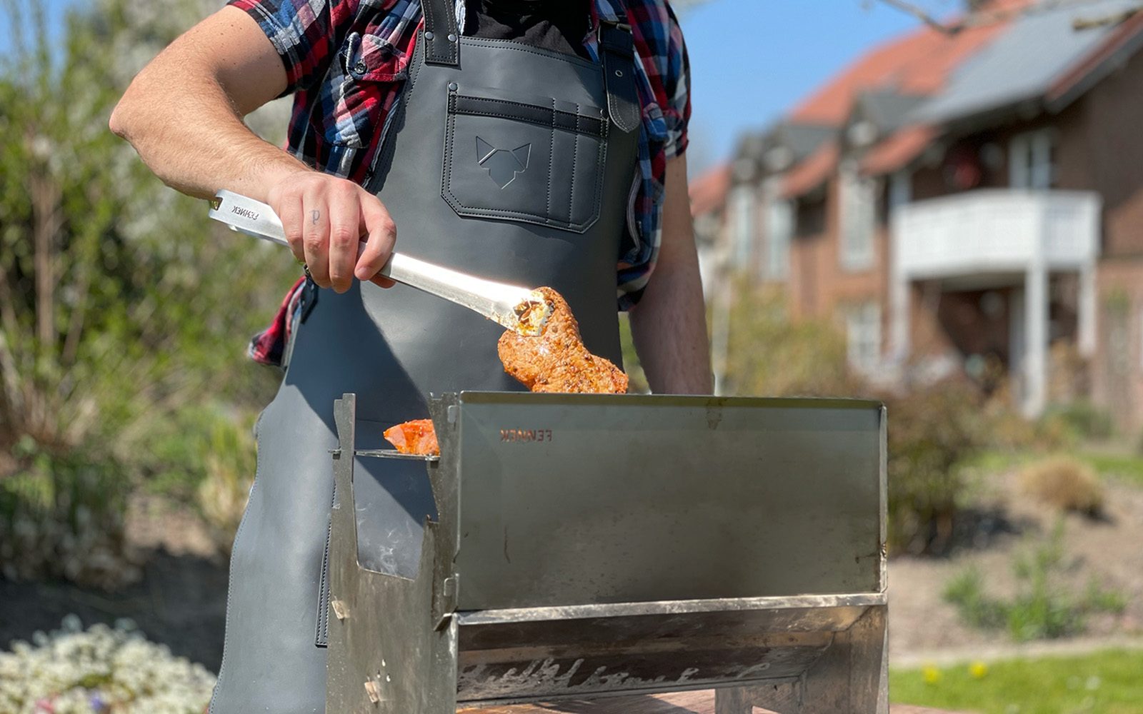 Grillschürze von einem Mann im Garten getragen im Einsatz an einem Holzkohlegrill, in der Hand eine Edelstahl-Grillzange die ein Stück Fleisch hält